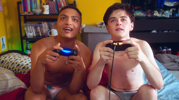 Two almost naked young men seated on a bed in a colourful room play a video game, from the short film Dungarees